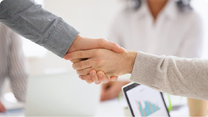 Side view of a handshake, with a blurred computer and another person in the background