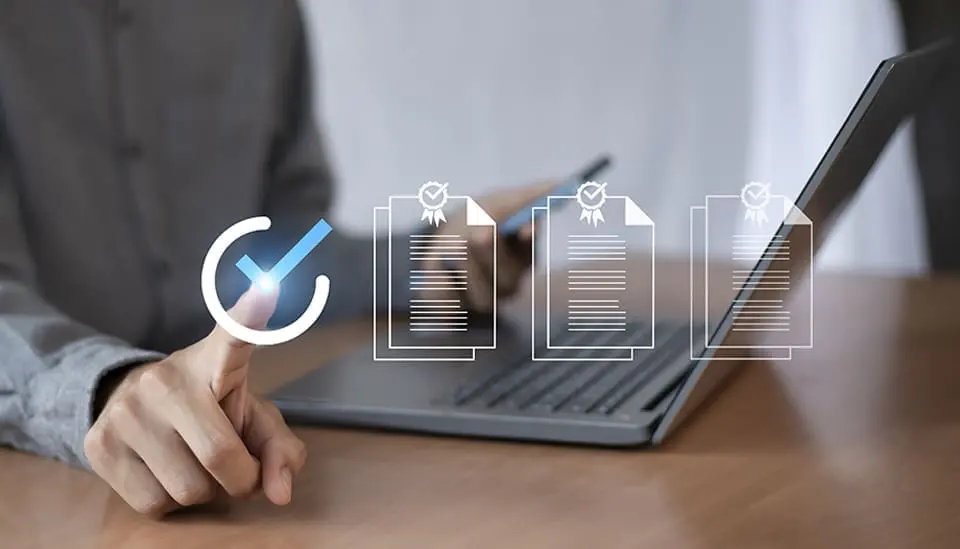 Una persona con traje utiliza un lápiz digital para marcar elementos en una lista de verificación virtual, con un portátil y una tableta sobre el escritorio.