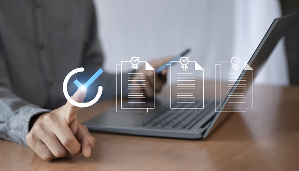 Una persona con traje utiliza un lápiz digital para marcar elementos en una lista de verificación virtual, con un portátil y una tableta sobre el escritorio.
